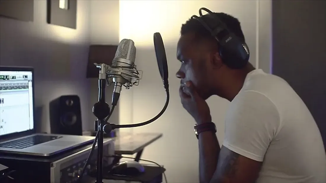 A person in headphones sings into a studio microphone beside a laptop running audio software.