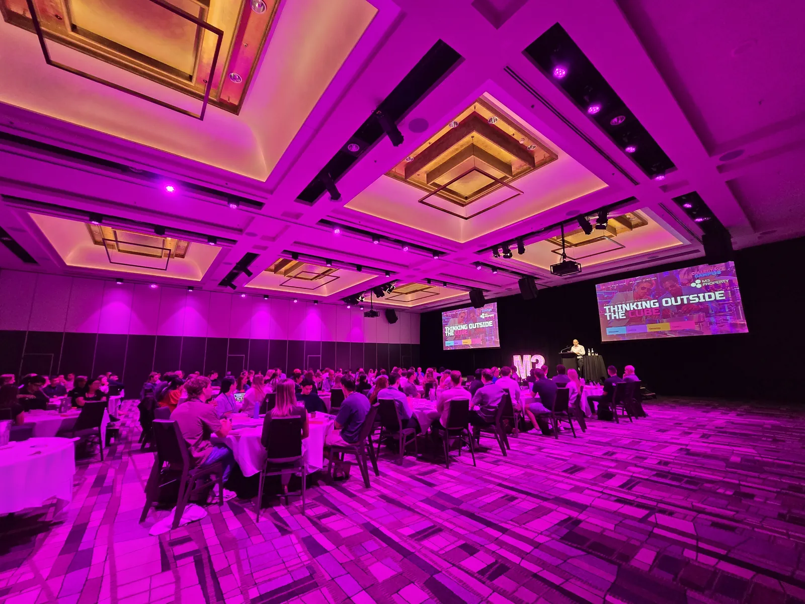 A wide view of the M3 Property ballroom with staff at tables and Thinking Outside the Cube on the screens